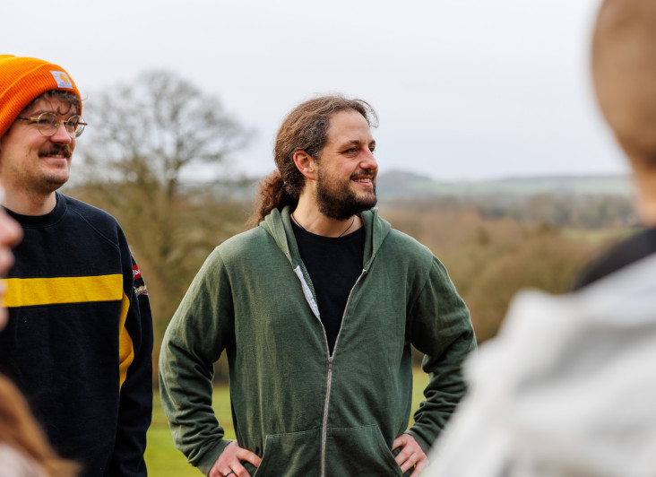 A group of people outdoors, with two men in the foreground. One man wears glasses and a black and yellow sweater, while the other has long hair and a green hoodie. The background features trees and rolling hills.