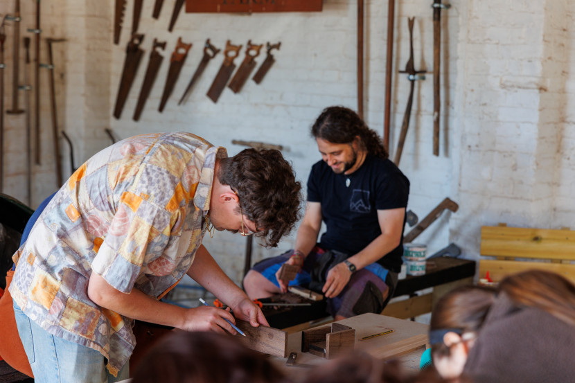 Two individuals engaged in woodworking in a well-lit workshop. One person, wearing a colorful shirt, is focused on measuring or marking a piece of wood, while another person, seated and dressed in dark clothing, is smiling and observing. Various hand tools, including saws, are visible on the wall behind them.