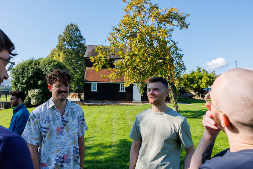 A group of four people stands in a grassy area with a house and trees in the background. They are engaged in conversation, smiling, and dressed casually, with one person wearing a patterned shirt and another with short hair. The sky is clear and blue, indicating a sunny day.