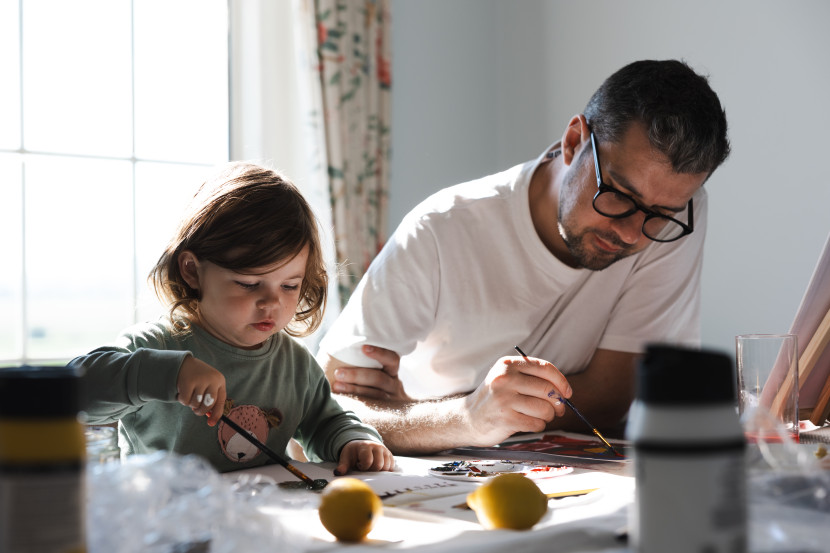 A father and his young child are sitting at a table, both engaged in painting. The child focuses intently on their artwork, while the father works on a piece of his own. Two lemons are placed on the table, and sunlight streams in through a nearby window.