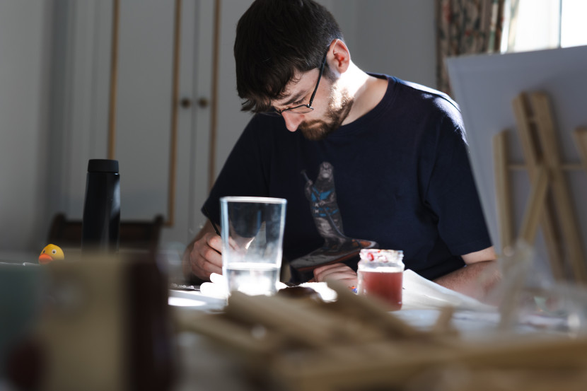 A man with glasses and a beard is focused on crafting or drawing at a table, surrounded by a glass of water, a candle, and various art supplies. Sunlight streams in, casting a warm glow on the scene.