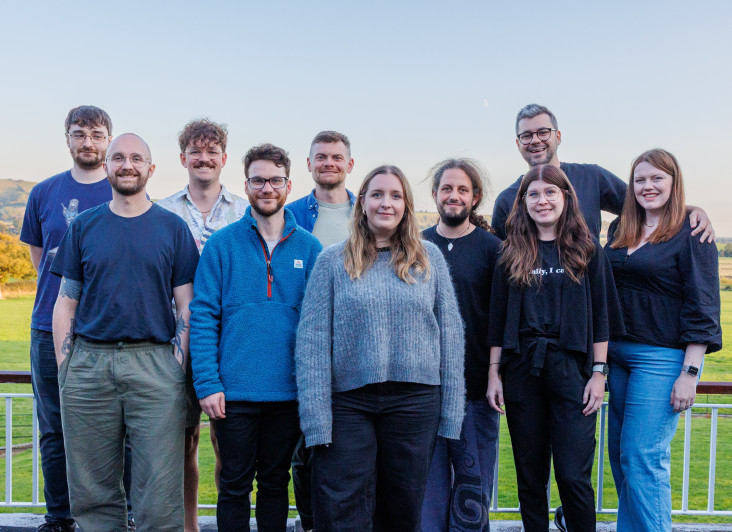 A group of eleven people stands outdoors on a balcony, smiling towards the camera with a scenic landscape in the background. They are dressed in casual clothing, with a mix of colors and styles, and display a range of hairstyles and expressions. The sky is clear with soft lighting.
