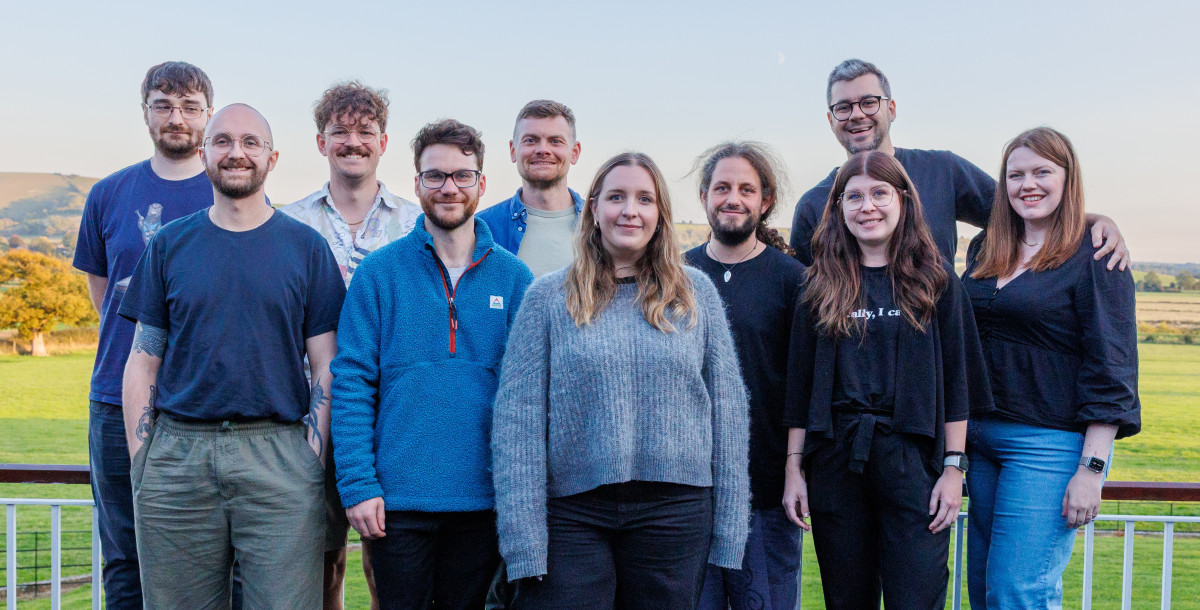 A group of eleven people stands outdoors on a balcony, smiling towards the camera with a scenic landscape in the background. They are dressed in casual clothing, with a mix of colors and styles, and display a range of hairstyles and expressions. The sky is clear with soft lighting.
