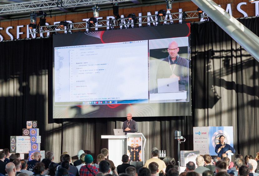 A speaker stands on stage in front of a large screen displaying coding on a laptop. An audience is seated in front, attentively watching. The venue features a modern design with banners and lighting.