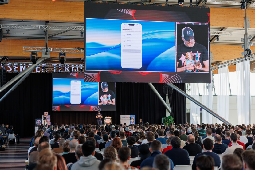 A speaker stands on stage holding a smartphone, presenting to a large audience. A large screen behind displays a mobile app interface against a blue background. The venue is bright, with many attendees seated, focused on the presentation.