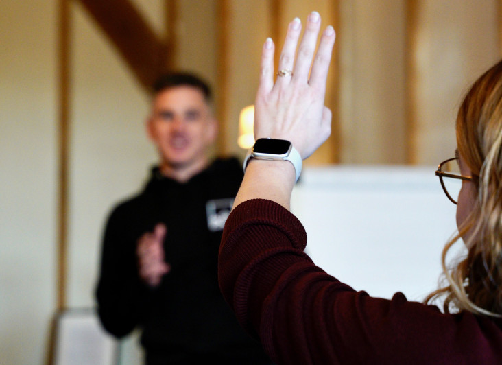 A person with a ring on their hand raises their arm in a questioning gesture, while a man wearing a black hoodie speaks in the background. The setting features wooden beams and a whiteboard.