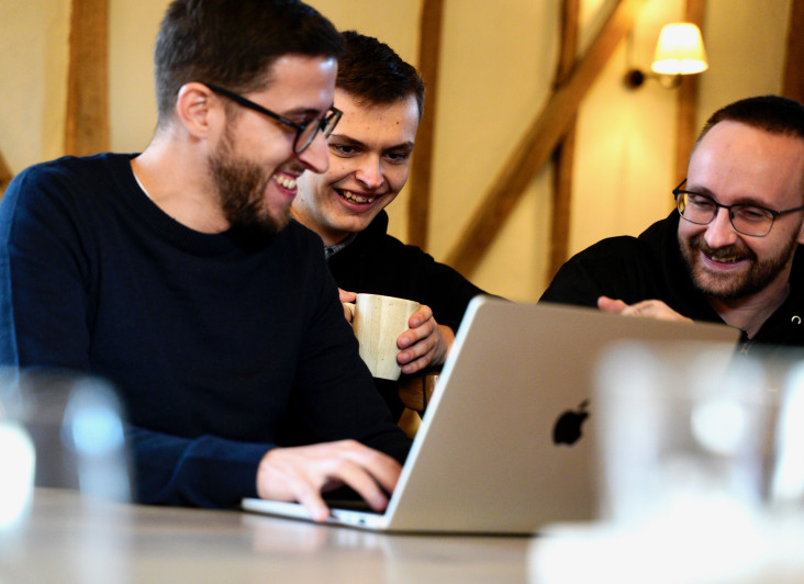 Three men are sitting at a table, collaborating over a laptop. One man, wearing glasses and a navy sweater, is typing on the laptop while the other two, one holding a mug, are smiling and looking at the screen. The setting has wooden beams and warm lighting.