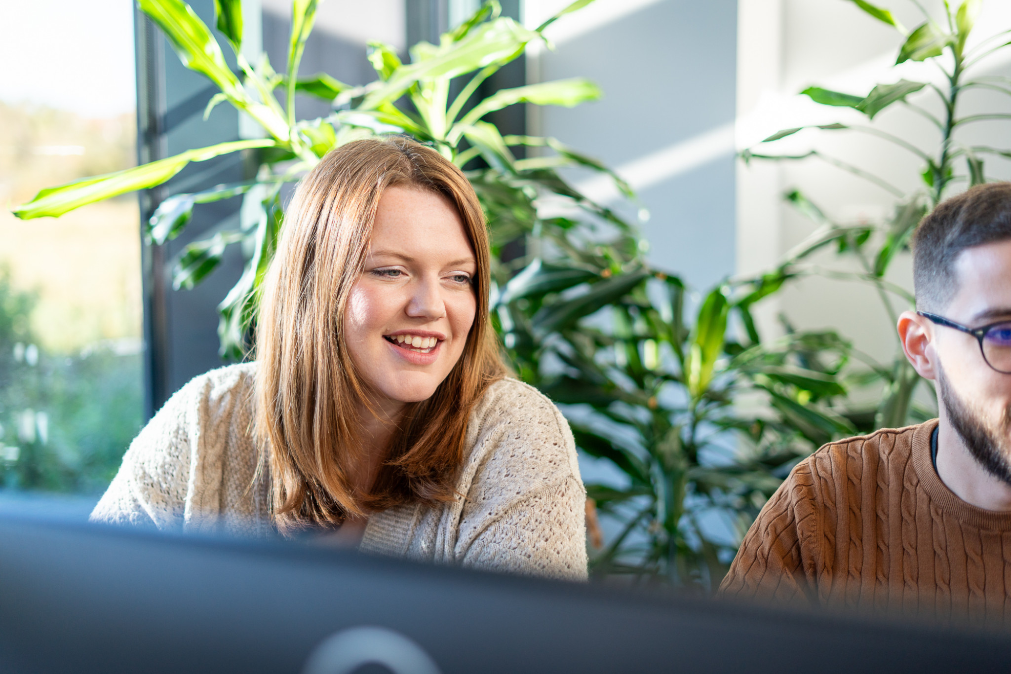 A woman with long brown hair smiles while sitting next to a man in a cozy sweater. They are in an office space filled with plants, with sunlight streaming in through the windows.