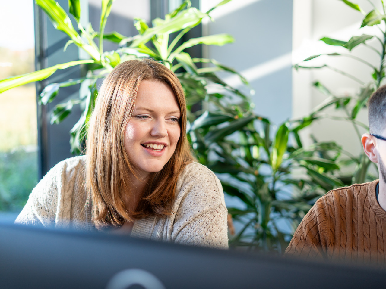 A woman with long brown hair smiles while sitting at a desk, engaged in conversation with a man beside her. Both are in a bright space filled with indoor plants. They appear to be working together on a project.