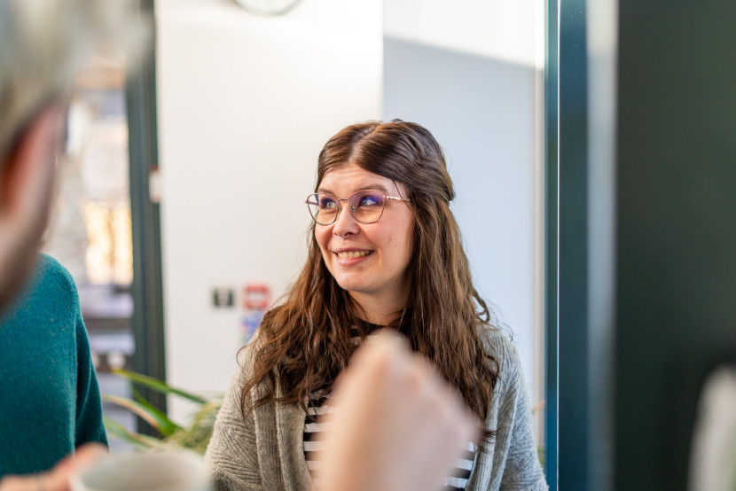 A woman with long, wavy brown hair wearing glasses smiles warmly while engaging in conversation. She is dressed in a gray sweater over a striped shirt and stands in a well-lit indoor setting with greenery in the background.