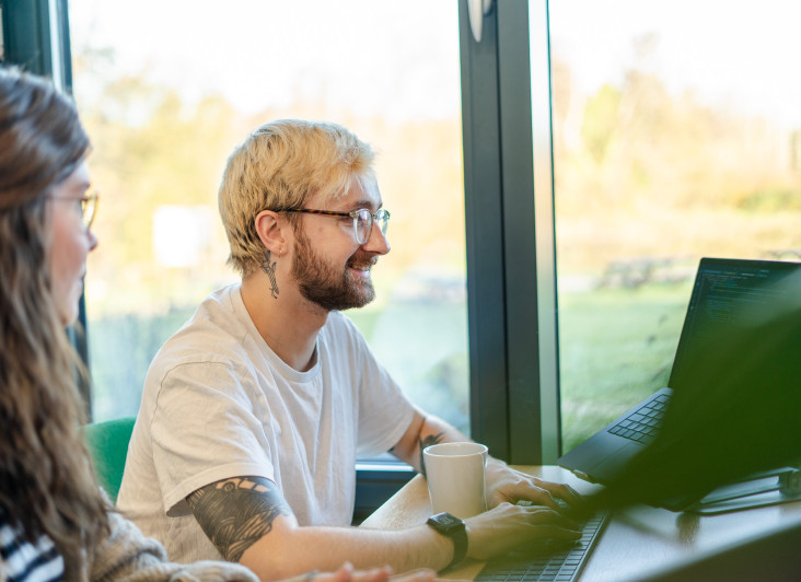 A young person with blonde hair and glasses smiles while working on a laptop at a table. Another individual, partially visible, sits beside them. Sunlight filters through large windows, illuminating a scenic outdoor view. A coffee cup rests on the table.