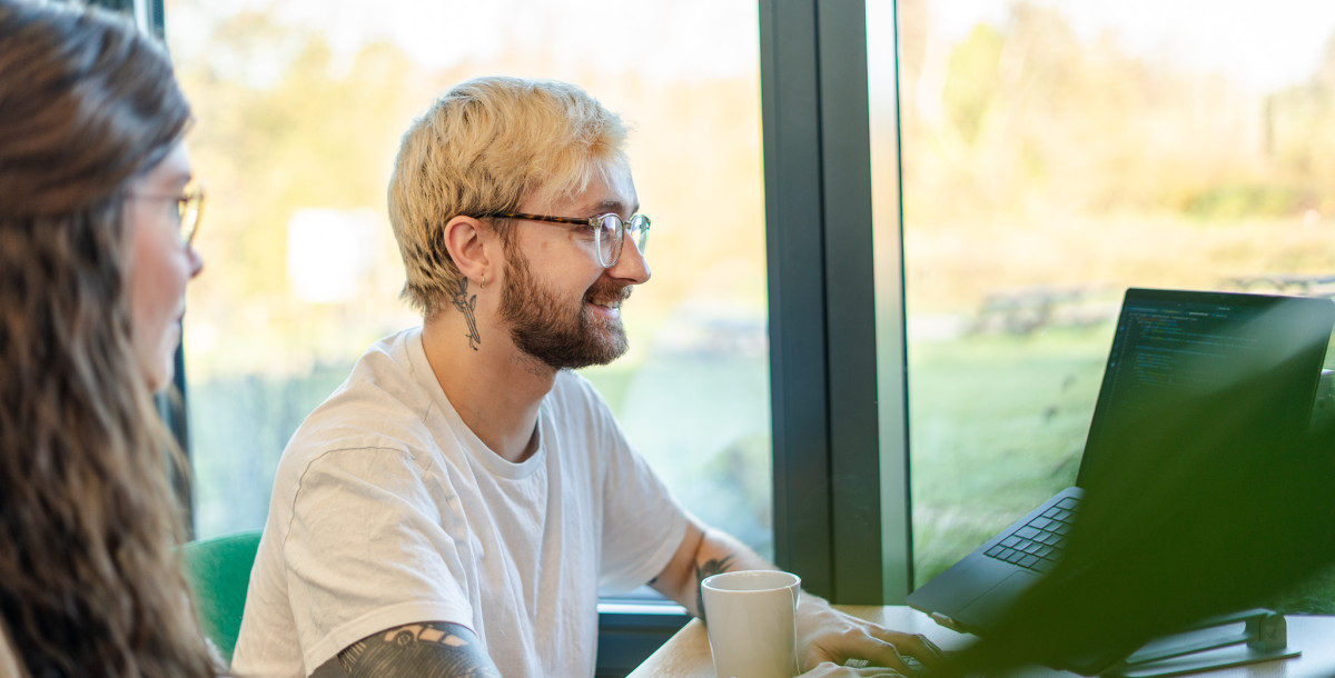 A young person with blonde hair and glasses smiles while working on a laptop at a table. Another individual, partially visible, sits beside them. Sunlight filters through large windows, illuminating a scenic outdoor view. A coffee cup rests on the table.