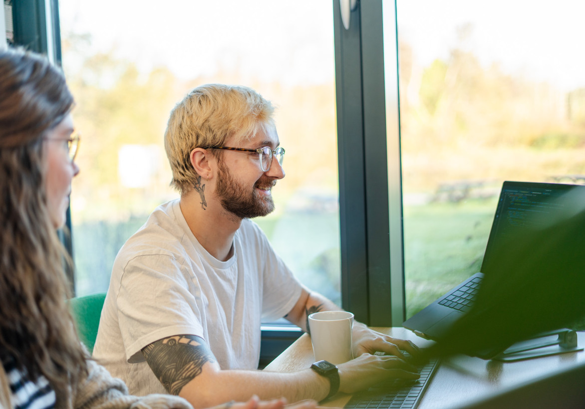 A young person with blonde hair and glasses smiles while working on a laptop at a table. Another individual, partially visible, sits beside them. Sunlight filters through large windows, illuminating a scenic outdoor view. A coffee cup rests on the table.