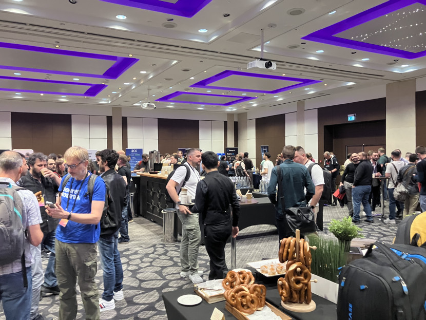 A brightly lit room full of groups of developers standing in small circles talking to each other, with tables of food in the foreground