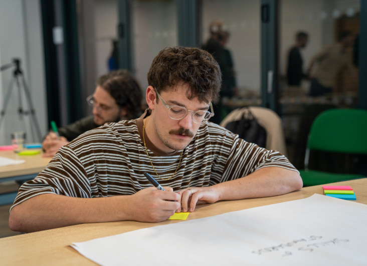 A person wearing glasses and a striped shirt writes on a yellow sticky note while sitting at a table. In the background, another person is also engaged in a similar activity. Various colored sticky notes are scattered on the table.
