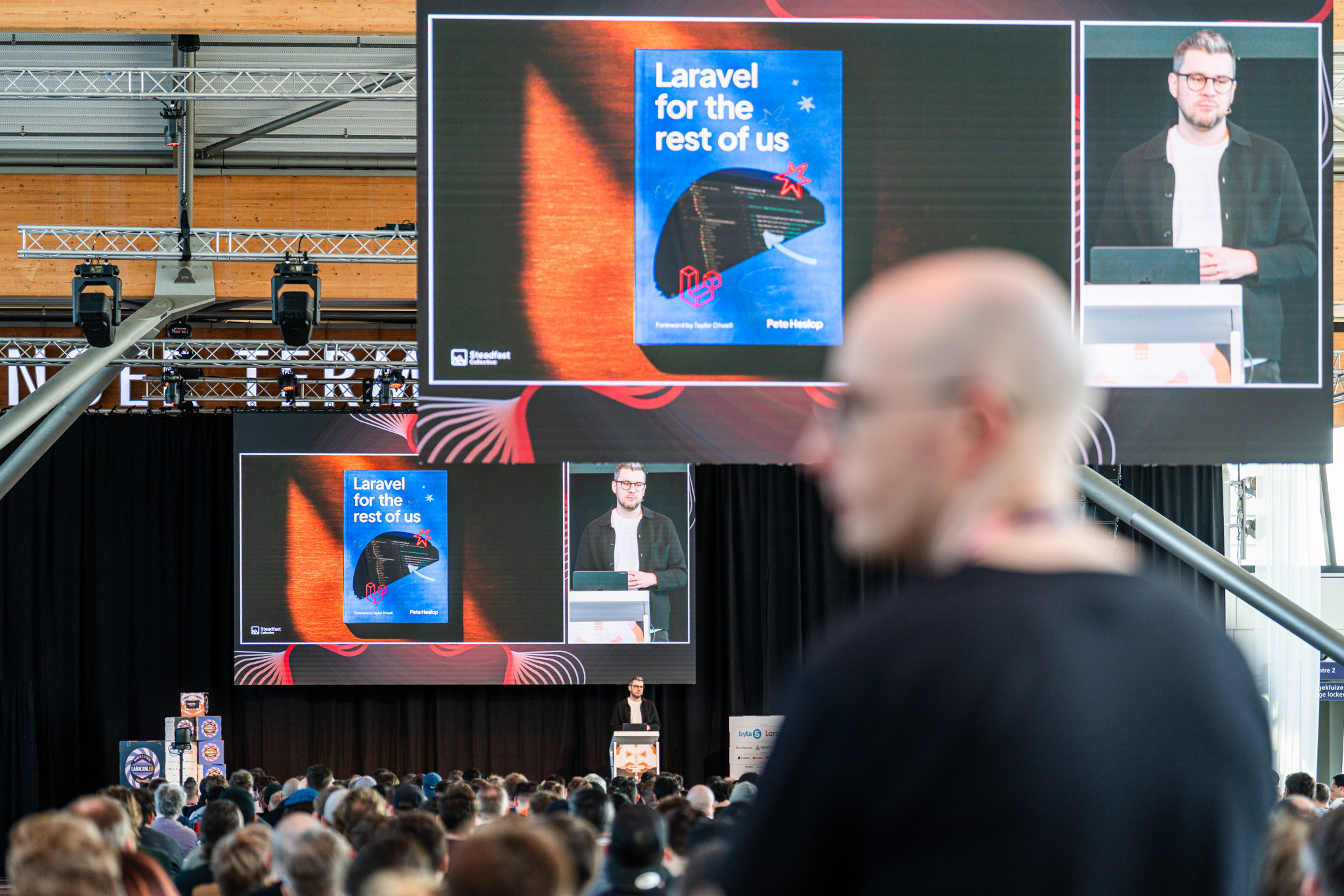 A speaker presents on stage at a conference, with a large screen displaying 
