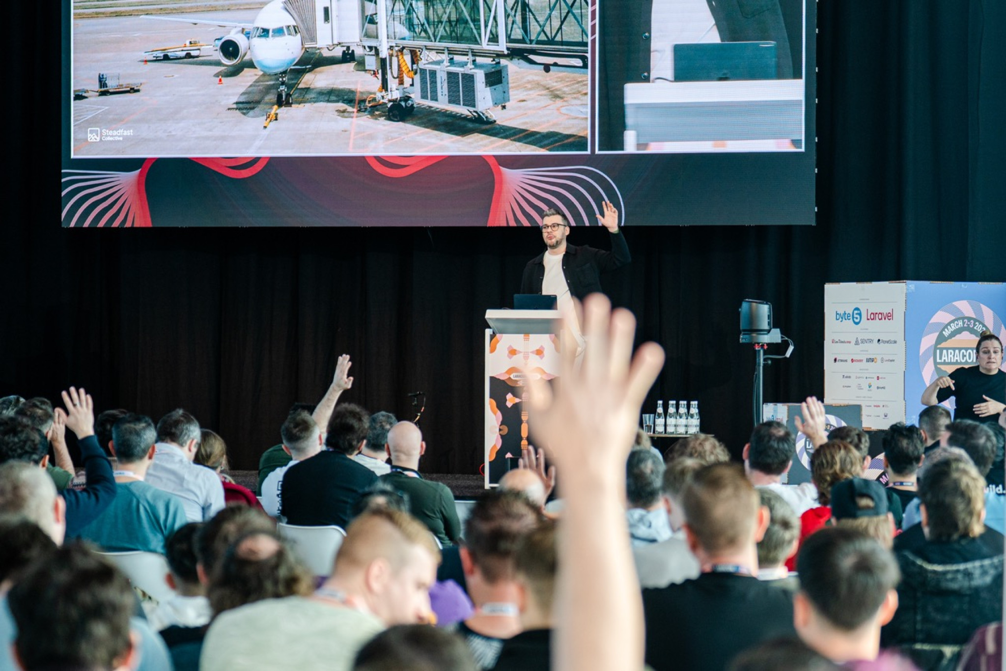 A speaker gestures while presenting on stage at a conference, with a large screen displaying an airplane outside a terminal in the background. Audience members raise their hands in response to a question.