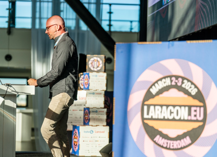 A speaker stands at a podium during a conference, engaged in a presentation. In the background, a large logo for Laracon EU features the event date and location. Bright lighting and a large screen are visible in the setting.
