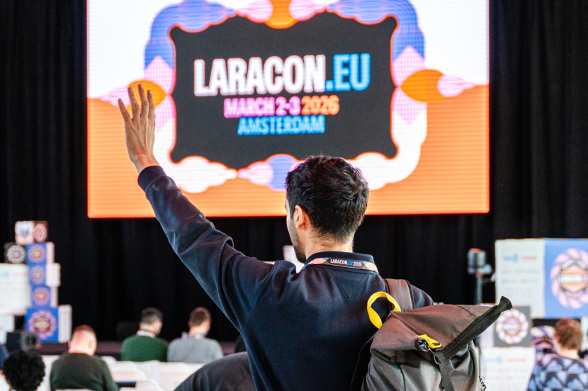 A person with short dark hair is waving their hand from the back of a conference audience. A large screen behind displays the text 