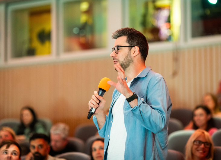 A speaker stands in front of an audience, holding a microphone with an orange top. He gestures with his hand while engaging the crowd. The setting appears to be a lecture or presentation space with others seated in the background.