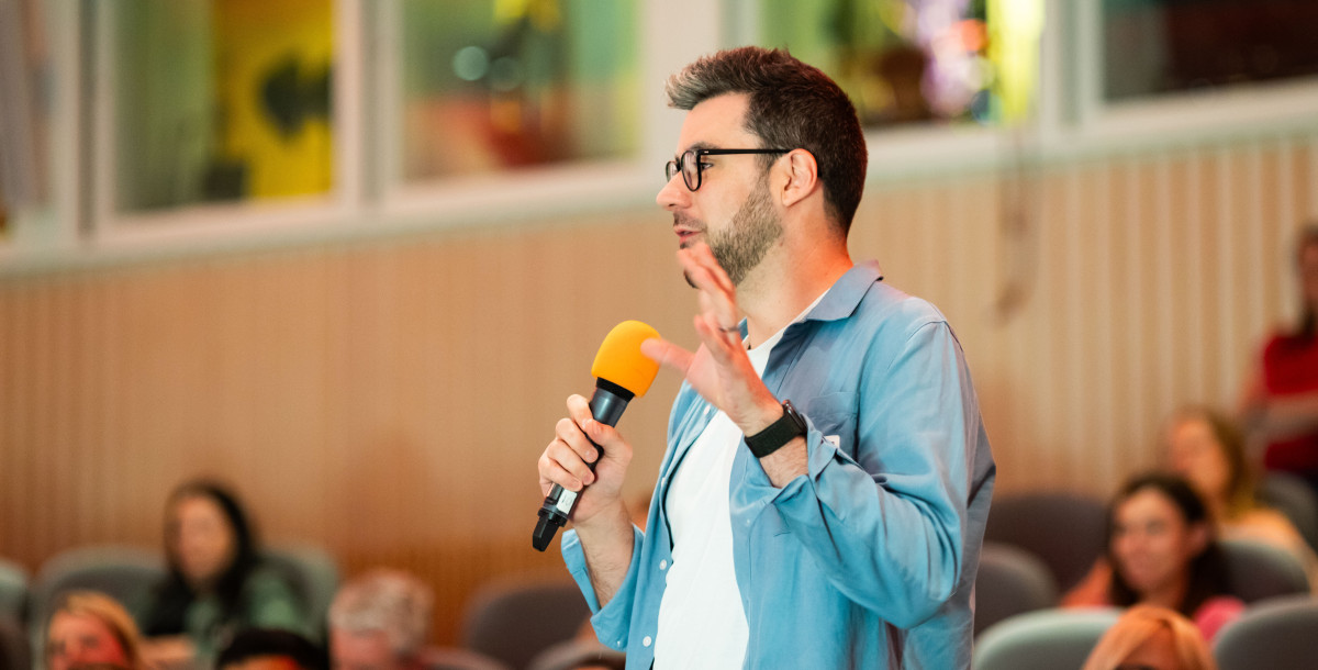A speaker stands in front of an audience, holding a microphone with an orange top. He gestures with his hand while engaging the crowd. The setting appears to be a lecture or presentation space with others seated in the background.