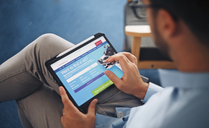 A person using a tablet to browse a website with a blue background and a search feature related to finding something, likely a service or product. They are seated with their legs crossed, wearing light-colored pants and a blue shirt.