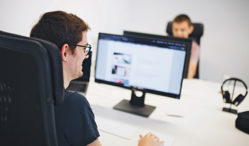 A person with glasses is sitting at a desk, working on a computer with a monitor displaying a website. In the background, another person is visible, also engaged in work. The setting appears to be a modern office.