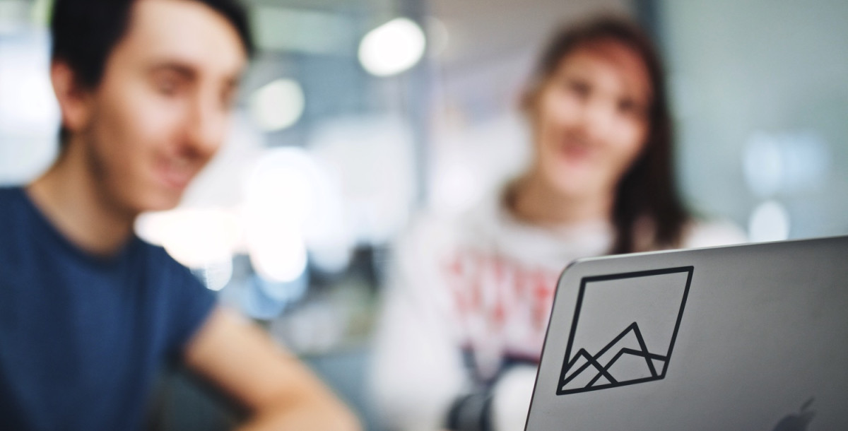A laptop with a mountain landscape sticker is in the foreground, while two people, blurred in the background, are engaged in conversation.